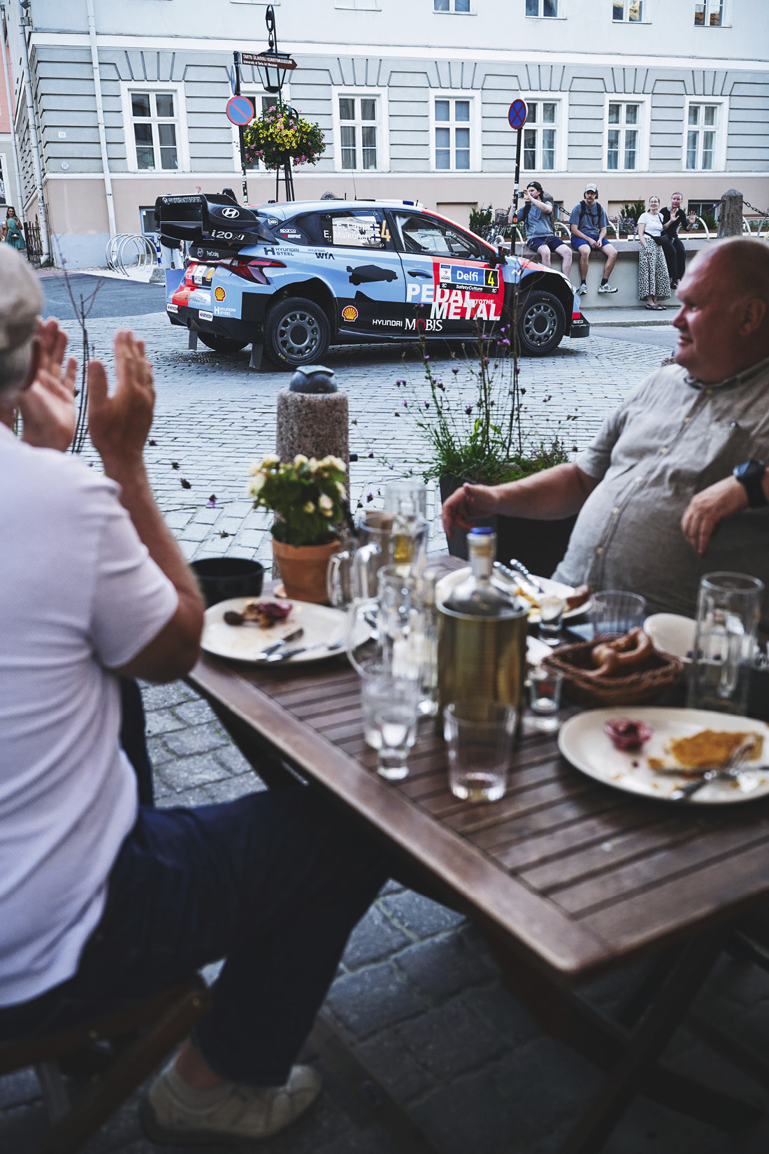 A rally car passes between two men dining in the city center