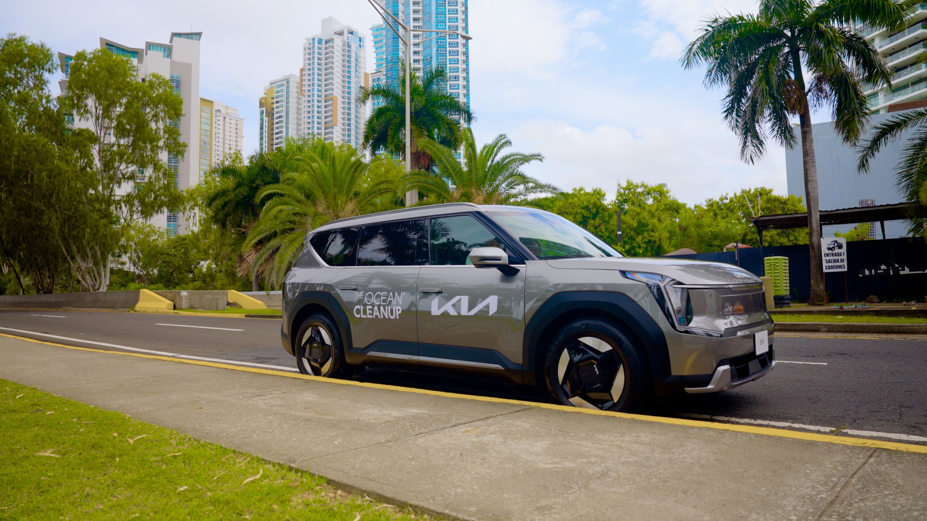 Grey Kia EV9 electric SUV branded with The Ocean Cleanup parked on a palm-lined urban street with modern high-rise buildings