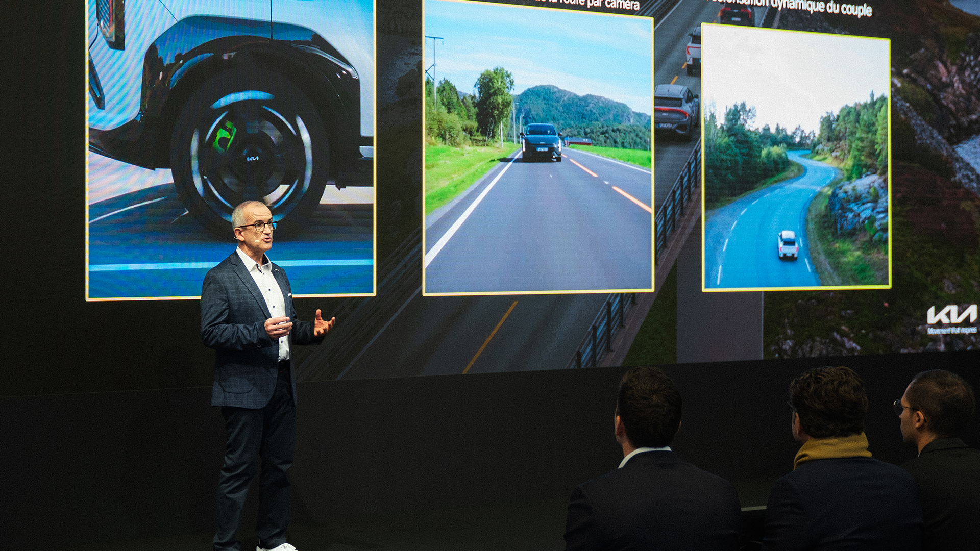 Kia presenter gestures beside driving-system visuals before audience