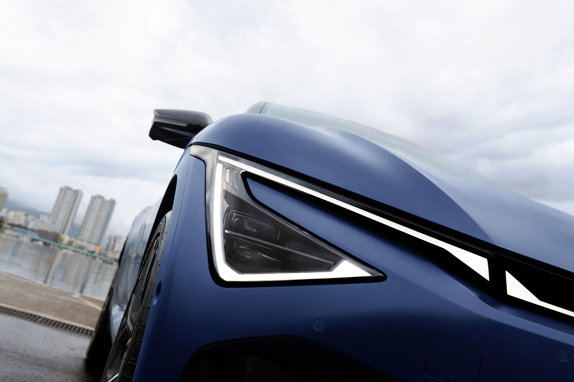 The sharp headlight of a blue EV6, viewed from a low angle against a waterfront promenade on a cloudy day