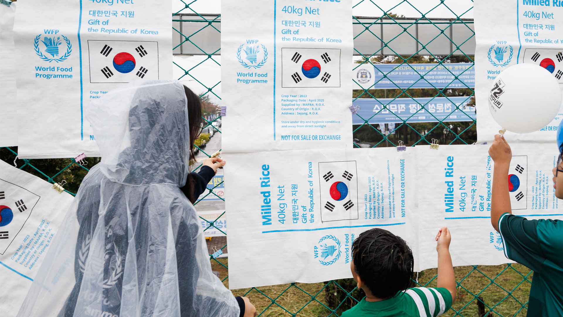 철망에 걸린 세계식량계획 쌀 포대 곁에 있는 초록 셔츠 아이들과 투명 우비를 입은 사람